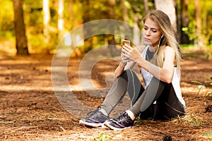 Beautiful blonde model in the middle of pines sitting using cellphone with headset. Listening to music