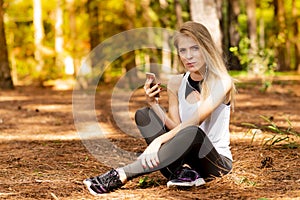 Beautiful blonde model in the middle of pines sitting using cellphone with headset. Listening to music