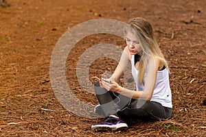 Beautiful blonde model sitting on dried pine leaves using cellphone and listening to music