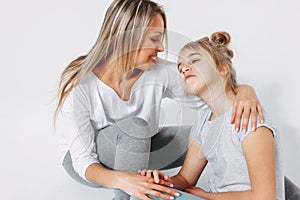 Beautiful blonde mom and daughter preteen in pajamas sitting and hugging isolated on white background