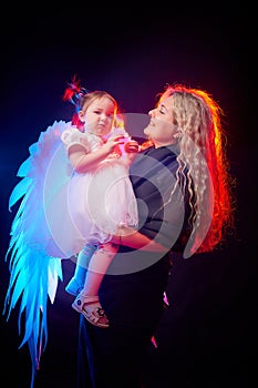A beautiful blonde mother with curly hair and cute small girl with white wings looking like a nice angels in photo shoot in studio