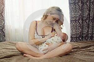 A beautiful blonde mother holds a newborn baby in her arms and gently touches his nose while sitting on the bed. Mom and