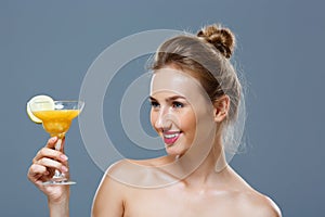 Beautiful blonde naked girl smiling, holding cocktail over grey background.