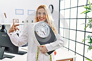 Beautiful blonde nutritionist woman holding weight machine to balance weight loss smiling happy pointing with hand and finger