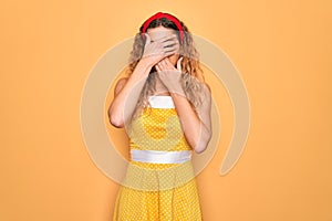 Beautiful blonde pin-up woman with blue eyes wearing diadem standing over yellow background Covering eyes and mouth with hands,