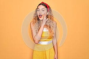 Beautiful blonde pin-up woman with blue eyes wearing diadem standing over yellow background hand on mouth telling secret rumor,
