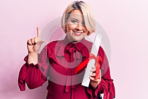 Beautiful blonde plus size woman holding graduated degree diploma over pink background smiling with an idea or question pointing