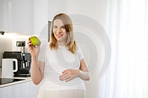 Beautiful blonde pregnant woman holding a pear and tough her belly standing in the kitchen