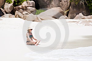Beautiful blonde pregnant woman in sun hat sits at white sand beach close to tropical green plants and granite rocks in Seychelles