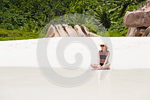 Beautiful blonde pregnant woman in sun hat sits at white sand beach close to tropical green plants and granite rocks in Seychelles