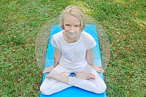 Beautiful blonde preteen girl in light clothing practicing yoga on a mat in the park. Healthy lifestyle.