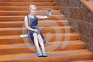 Beautiful blonde preteen girl using a phone, makes the photo a front camera, sitting on a rusty staircase in a modern building