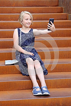 Beautiful blonde preteen girl using a phone, makes the photo a front camera, sitting on a rusty staircase in a modern building