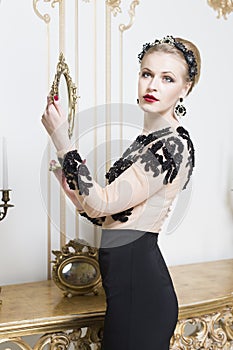 Beautiful blonde royal woman standing near retro table in gorgeous luxury dress looking in front of herself. Indoor