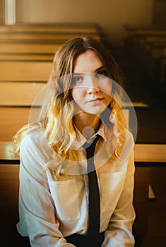 A beautiful blonde schoolgirl dressed in a white shirt and black tie is sitting alone at a school desk in a classroom. Educational