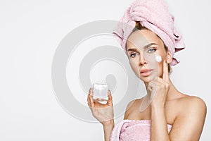 Beautiful blonde woman model holding a cream in her hand and creaming her face with towel on her head