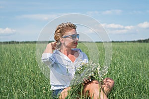 Beautiful blonde short-haired woman in sunglasses sits on a green meadow on a summer day and holds field daisies