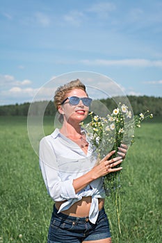 Beautiful blonde short-haired woman in sunglasses sits on a green meadow on a summer day and holds field daisies