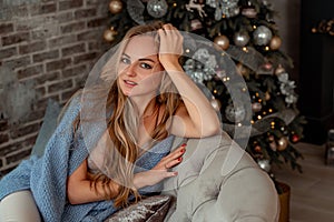 A beautiful blonde is sitting on a sofa in a room with Christmas decorations. Winter mood, cozy home, against the