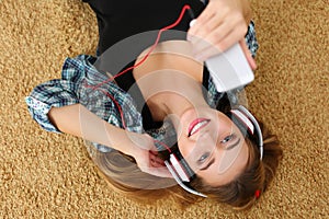 Beautiful blonde smiling woman lying on carpet floor wearing headphones, holding cell phone and listening music portrait. Modern