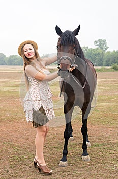 Beautiful blonde in a straw hat and summer dress, photographed with a horse