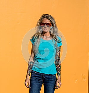 Beautiful blonde in sunglasses look at the camera. Portrait on the background of bright orange wall. Modern hipster girl