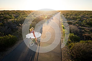 Beautiful blonde surfer girl on her way to the beach on her bicycle with her surfboard.