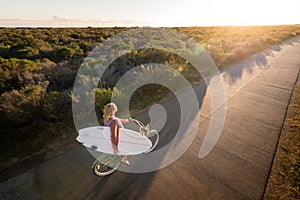 Beautiful blonde surfer girl on her way to the beach on her bicycle with her surfboard.