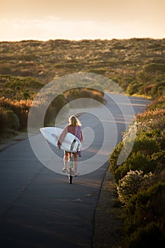 Beautiful blonde surfer girl on her way to the beach on her bicycle with her surfboard.