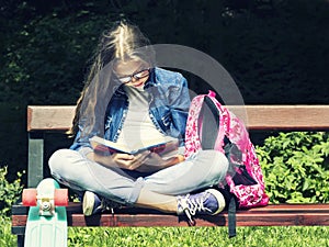 Beautiful blonde teen girl in jeans shirt reading a book on the bench with a backpack and skateboard in the park