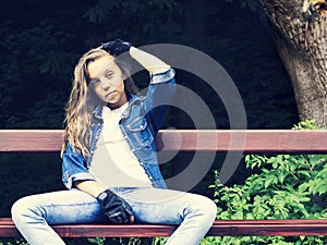 Beautiful blonde teen girl in jeans shirt, sitting on bench with backpack and skateboard in park