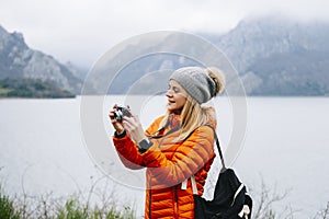 beautiful blonde tourist woman, with a wool hat, backpack and orange coat, uses the camera in a landscape with a lake and