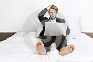 Beautiful blonde unshaved businessman lying in bed, working on laptop computer, holding hand on head with shocked