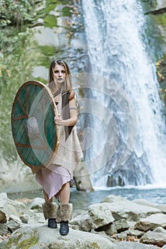 Beautiful blonde viking woman with a shield and a sword near a waterfall