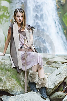 Beautiful blonde viking woman with a shield and a sword near a waterfall