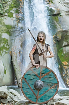 Beautiful blonde viking woman with a shield and a sword near a waterfall