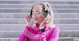 Beautiful blonde in wireless headphone listening to music and dancing on urban stairs in park, close up