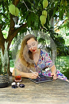 Beautiful blonde woman artist and illustrator drawing, sitting by the table with electronical tablet and stylus