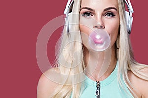 Beautiful blonde woman blowing pink bubble gum. Girl listen music in headphones. Fashion and lifestyle portrait.