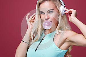Beautiful blonde woman blowing pink bubble gum. Girl listen music in headphones. Fashion and lifestyle portrait.
