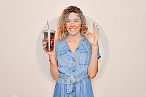 Beautiful blonde woman with blue eyes drinking cola beverage using straw to refreshment doing ok sign with fingers, excellent