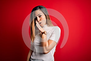Beautiful blonde woman with blue eyes wearing casual white t-shirt over red background hand on mouth telling secret rumor,
