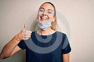 Beautiful blonde woman with blue eyes wearing medical mask over white background doing happy thumbs up gesture with hand