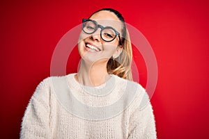 Beautiful blonde woman with blue eyes wearing sweater and glasses over red background happy face smiling with crossed arms looking