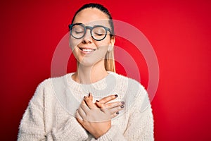 Beautiful blonde woman with blue eyes wearing sweater and glasses over red background smiling with hands on chest with closed eyes