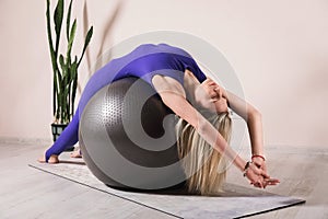 Beautiful blonde woman in blue sportswear practicing yoga performing bridge pose, Bandhasana on fitball in the room