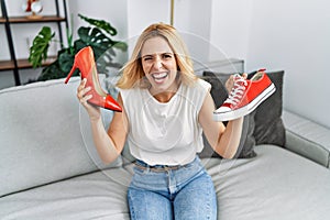 Beautiful blonde woman choosing high heel shoes and sneakers at home angry and mad screaming frustrated and furious, shouting with