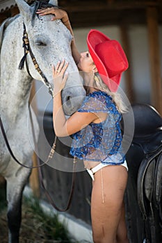 Beautiful blonde woman with curly hair, hat and horse. Portrait of a girl with white bikini blue transparent blouse and her horse