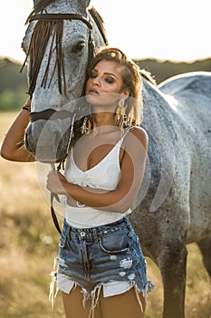 Beautiful blonde woman with curly hair with white hat and horse. Portrait of a girl with denim and her horse.