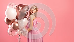 Beautiful blonde woman in cute dress holding balloons, pursing her lips for kiss, being playful on pink background